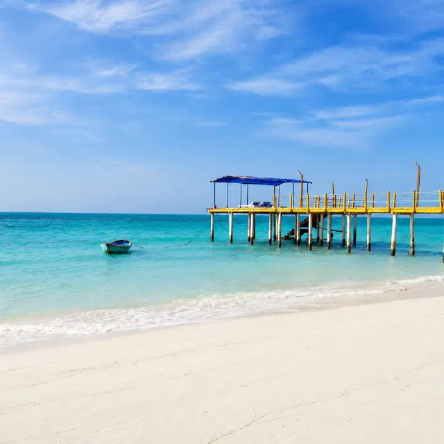 Bright tropical Agatti beach with turquoise water, white sand, a small boat, and a yellow pier extending into the sea.