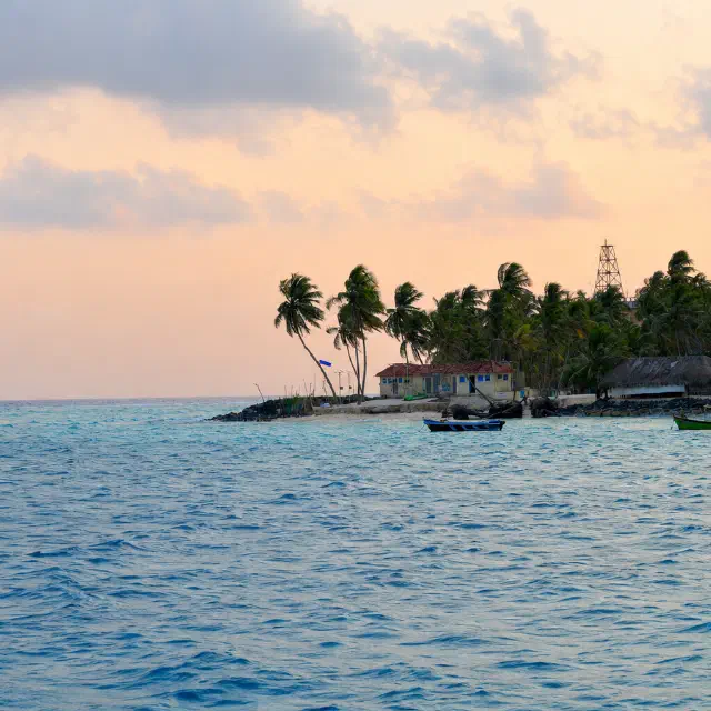 Palm-covered Kavaratti island north coastline with small boats on blue water under a soft pastel sky near sunset or dawn.