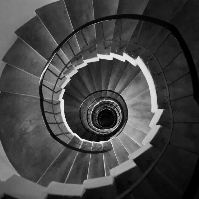 Black-and-white photo looking down a spiral staircase with curved railings forming a hypnotic circular pattern.