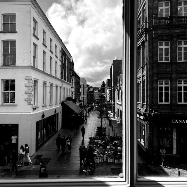 Photo of a narrow city street seen through a window, with pedestrians below, tall buildings on both sides, and dramatic clouds above.