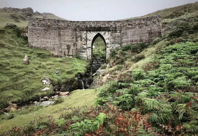 Old stone archway standing among green hills and ferns, with a small stream running beneath it.