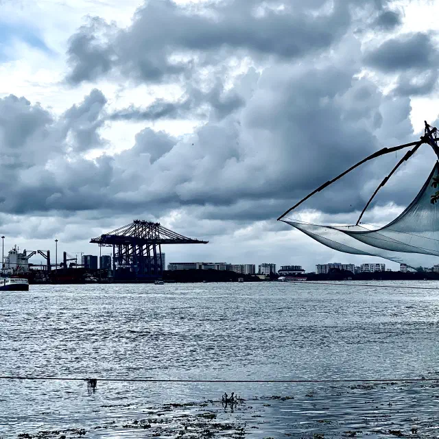 Black-and-white photo of a harbor with dramatic storm clouds, water, a distant boat, port cranes, and a Chinese fishing net on the right.