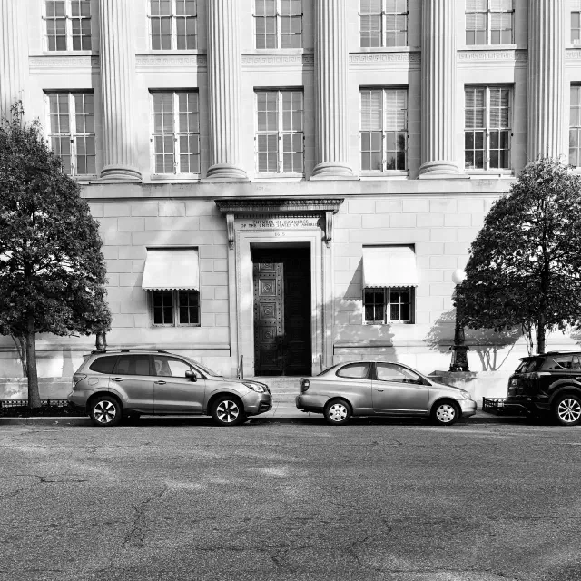 Black-and-white photo of a grand building facade with tall columns, windows, trees, and parked cars outside.