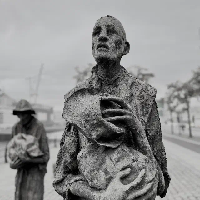 Black-and-white close-up of an Irish Famine memorial sculpture showing a gaunt figure holding a bundle, with another blurred figure in the background