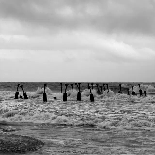 Black-and-white photo of waves crashing around old sea pillars at Kozhikode Beach under a cloudy sky.